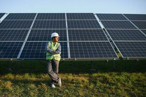 Porträt von jung indisch Mann Techniker tragen Weiß schwer Hut Stehen in der Nähe von Solar- Paneele gegen Blau Himmel. industriell Arbeiter Solar- System Installation foto