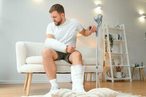 Wiederherstellung nach physisch Verletzung im Unfall beim Zuhause traurig unglücklich jung Mann mit gebrochen Bein, verletzt Arm foto