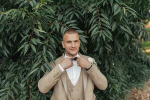 Hochzeit Foto auf ein Grün Hintergrund. das Bräutigam steht in der Nähe von das Grün Gebüsch, passt sich an das Schmetterling und sieht aus in das Linse. Porträt von das Bräutigam