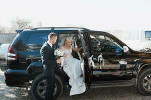 Luxus Hochzeit Fotografie. elegant Hochzeit Paar, das Bräutigam im ein schwarz passen und das Braut im ein Hochzeit Kleid mit ein Strauß von Blumen im das Auto, das Bräutigam Küsse das Hand von das Braut foto