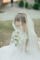 lockig blond Braut im ein Weiß Kleid, bedeckt mit ein Schleier, posiert zum das Kamera mit ein Strauß von Rosen. Porträt von das Braut. schön bilden und Haar. Hochzeit im Natur foto