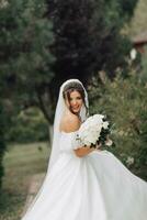 Hochzeit Foto. das Braut im ein voluminös Weiß Kleid und ein lange Schleier, lächelnd, wirbelnd mit ein Strauß von Weiß Rosen, halten ihr Kleid. Porträt von das Braut. schön bilden und Haar. foto