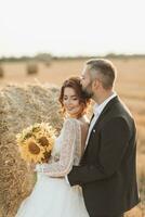 Hochzeit Porträt von das Braut und Bräutigam. das Bräutigam Umarmungen das Braut von hinter, Nächster zu ein Ballen von Heu. Seite Sicht. rothaarig Braut im ein lange Kleid mit ein Strauß von Sonnenblumen. stilvoll Bräutigam. Sommer- foto