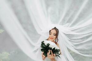 lockig Brünette Braut im ein Weiß Kleid, bedeckt mit ein Schleier, posiert zum das Kamera mit ein Strauß von Rosen. Porträt von das Braut. schön bilden und Haar. Hochzeit im Natur foto