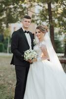 Hochzeit Porträt. das Bräutigam im ein schwarz passen und das blond Braut sind Stehen, umarmen, posieren halten ein Strauß unter ein Baum. Foto Session im Natur. schön Haar und bilden