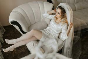 blond Braut tragen ein Satin- Kleid und Lügen auf ein Couch, posieren und zeigen aus ihr Tiara und Schleier. schön Haar und bilden, öffnen Büste. Hochzeit Porträt. aufrichtig Lächeln foto