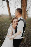 Hochzeit Foto. das Braut und Bräutigam sind Gehen im das Wald. das Bräutigam Umarmungen das Schultern von seine geliebte. lange Hochzeit Kleid. ein Paar im Liebe unter hoch Bäume. Herbst Sonnenlicht. foto