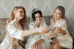 ein Brünette Braut und ihr blond freunde sind Sitzung auf das Sofa im das Braut Zimmer, tragen Bademäntel, lächelnd und Klirren Champagner Brille. Hochzeit Porträt Hochzeit Foto im ein Licht Schlüssel