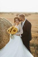 Hochzeit Porträt von das Braut und Bräutigam. das Bräutigam Umarmungen das Braut von hinter, Nächster zu ein Ballen von Heu. Seite Sicht. rothaarig Braut im ein lange Kleid mit ein Strauß von Sonnenblumen. stilvoll Bräutigam. Sommer- foto