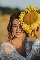 Hochzeit Porträt. ein rothaarig Braut im ein Weiß Kleid Abdeckungen ihr Auge mit ein Sonnenblume, lächelt und sieht aus beim das Kamera. schön Locken. aufrichtig lächeln. elegant Kleid. foto