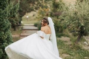 Hochzeit Foto. das Braut im ein voluminös Weiß Kleid und ein lange Schleier, lächelnd, wirbelnd mit ein Strauß von Weiß Rosen, halten ihr Kleid. Porträt von das Braut. schön bilden und Haar. foto