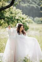 ein schön Braut mit lange lockig Haar hält ihr zart Kleid und bewundert es im das Garten. Morgen von das Braut. natürlich bilden foto