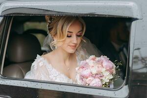 ein blond Braut sitzt im ein schwarz Auto auf ihr Hochzeit Tag mit ein Strauß. Porträt von das Braut. schön bilden und Haar. großartig Weiß Spitze Kleid foto