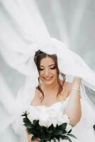 lockig Brünette Braut im ein Weiß Kleid, bedeckt mit ein Schleier, posiert zum das Kamera mit ein Strauß von Rosen. Porträt von das Braut. schön bilden und Haar. Hochzeit im Natur foto