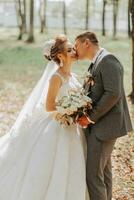 modisch Bräutigam und süß blond Braut im ein Weiß Kleid mit ein Krone, ein Strauß sind umarmen, Lachen im das Park, Garten, Wald draußen. Hochzeit Fotografie, Porträt von lächelnd Jungvermählten. foto