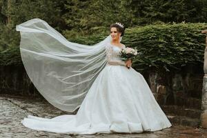 voll Länge Seite Aussicht von einer schön sinnlich jung Brünette Braut im lange Weiß Hochzeit Kleid und Schleier Stehen im Park halten Strauß draußen auf natürlich Hintergrund, horizontal Bild foto