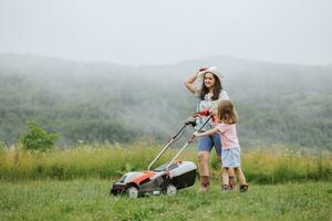 ein Frau im Stiefel mit ihr Kind im das bilden von ein Spiel mäht das Gras mit ein Rasenmäher im das Garten gegen das Hintergrund von Berge und Nebel, Garten Werkzeuge Konzept, arbeiten, Natur. foto