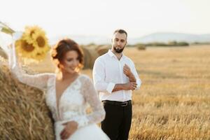 Hochzeit Porträt von das Braut und Bräutigam. das Bräutigam, reißen seine Shirt, steht hinter das Braut, in der Nähe von ein Ballen von Heu. rothaarig Braut im ein lange Kleid mit ein Strauß von Sonnenblumen. stilvoll Bräutigam. Sommer- foto