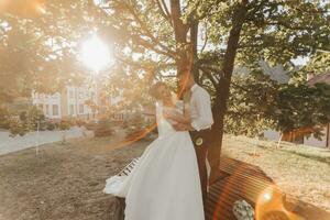 Hochzeit Porträt. ein Bräutigam im ein schwarz passen und ein blond Braut sind Stehen, umarmen und küssen unter ein Baum. Foto Session im Natur. ein schön Strahl von das Sonne im das Foto. schön Haar und bilden