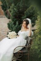 Porträt von das Braut im Natur. Brünette Braut im ein Weiß lange Kleid, halten ein Strauß von Weiß Rosen, Sitzung auf ein Bank, posieren, suchen runter, gegen ein Baum Hintergrund. lockig Haar foto