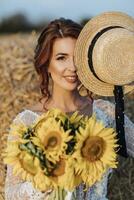 Hochzeit Porträt. ein rothaarig Braut im ein Weiß Kleid hält ein Strauß von Sonnenblumen und Abdeckungen ihr Gesicht mit ein stilvoll Hut, lächelt und sieht aus beim das Kamera. schön Locken. aufrichtig lächeln. foto