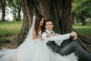 ein schön Braut mit lange lockig Haar im ein schick Kleid Umarmungen das Bräutigam, lächelt, suchen in das Linse unter ein groß Baum. Porträt von das Braut und Bräutigam. Frühling Hochzeit. natürlich bilden foto