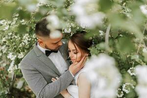 das Braut im ein Weiß Abend Kleid posiert mit das Bräutigam mit ein schön Frisur, das Bräutigam sanft Wraps das Braut und lehnt sich gegen ihr gegen das Hintergrund von Frühling Blumen. foto