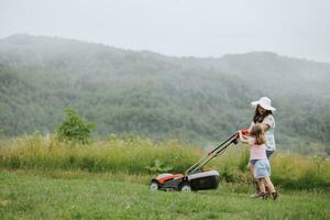 ein Frau im Stiefel mit ihr Kind im das bilden von ein Spiel mäht das Gras mit ein Rasenmäher im das Garten gegen das Hintergrund von Berge und Nebel, Garten Werkzeuge Konzept foto