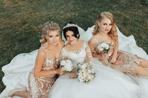 ein Brünette Braut im ein Weiß elegant Kleid mit ein Krone und ihr blond freunde im Gold Kleider Pose mit Blumensträuße während Sitzung auf das Gras. Hochzeit Porträt im Natur, Hochzeit Foto im ein Licht Ton.