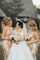 ein Brünette Braut im ein Weiß elegant Kleid mit ein Krone und ihr blond freunde im Gold Kleider Pose mit Blumensträuße. Hochzeit Porträt im Natur, Hochzeit Foto im ein Licht Ton.