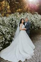 glücklich jung Paar, Braut mit lange lockig Haar im Weiß Kleid mit lange Zug in der Nähe von Schloss im schön Blumen. schön Mädchen im das Park. schön Sonnenlicht. Hochzeit Foto schießen