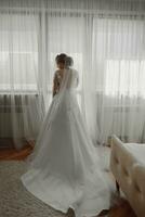 Vorbereitung zum das Hochzeit. schön jung Braut im Weiß Hochzeit Kleid drinnen. Luxus Modell- sieht aus beim Hochzeit Schuhe, beim Zuhause im Studio Zimmer mit groß Fenster. das Mädchen zeigt an ein Foto von das zurück