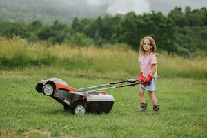 ein Kind im Stiefel im das bilden von ein Spiel mäht Gras mit ein Rasenmäher im das Hof gegen das Hintergrund von Berge und Nebel, das Konzept von Garten Werkzeuge foto