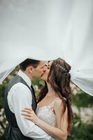 glücklich jung Paar. Hochzeit Porträt. das Braut und Bräutigam zärtlich Kuss unter ein Schleier gegen das Hintergrund von ein Blühen Busch. Hochzeit Strauß. Frühling Hochzeit foto