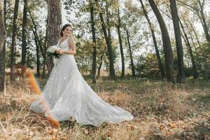 Hochzeit Foto im Natur. das Braut ist Stehen im das Wald. das Braut im ein schön Kleid mit ein lange Zug, halten ihr Strauß von Weiß Rosen, lächelnd Mit freundlichen Grüßen beim das Kamera. Porträt