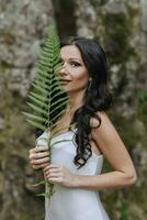 ein Mädchen mit lange schwarz lockig Haar, Stehen im ein Weiß Seide Kleid im ein Frühling Wald gegen ein groß Baum, halten ein Farn Ast im ihr Hände foto