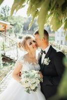 Hochzeit Porträt. das Bräutigam im ein schwarz passen und das blond Braut Stand durch ein Stein Mauer unter ein Baum, das Bräutigam Küsse das Braut Hand. Foto Session im Natur. schön Haar und bilden