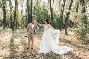 Hochzeit Foto. das Braut und Bräutigam sind Gehen im das Wald. das Bräutigam hält ein Strauß und Küsse das Hand von seine geliebte, und sie schön wirft oben ihr lange Schleier. Paar im Liebe. Sommer- Licht. foto