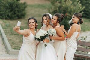 Hochzeit Foto im Natur. ein Brünette Braut im ein lange Weiß Kleid und ihr freunde im nackt Kleider sind lächelnd und nehmen ein Selfie auf ihr Telefon, halten ihr Gypsophila Blumensträuße. jung Frauen. Emotionen