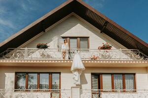 Weiß Hochzeit Kleid hängend auf Balkon Geländer, breit Schuss von Haus im Vorderseite und Mädchen auf Balkon. Hochzeit Einzelheiten, modern Hochzeit Kleid mit lange Zug, lange Ärmel und öffnen zurück foto