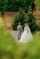 Hochzeit im Natur. das Braut steht im Vorderseite von das Bräutigam und sieht aus Über seine Schulter gegen ein Hintergrund von Bäume und ein hölzern Hütte. stilvoll Bräutigam. schön Braut. foto