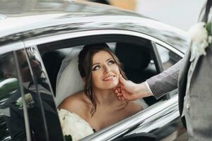 Hochzeit Foto. das Braut sitzt im ein schwarz Auto mit ein lächeln, das Bräutigam sanft hält das Braut kinn. Aussicht durch das Auto Fenster. schön bilden und Haar. ein luxuriös Weiß Kleid foto