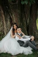 ein schön Braut mit lange lockig Haar im ein schick Kleid Umarmungen das Bräutigam, lächelt, suchen in das Linse unter ein groß Baum. Porträt von das Braut und Bräutigam. Frühling Hochzeit. natürlich bilden foto