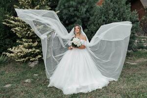 ein lockiges Haar Brünette Braut im ein Weiß Kleid und Schleier, fließend schön im das Luft, steht im Vorderseite von ihr stilvoll Bräutigam. Porträt von das Braut und Bräutigam. Hochzeit im Natur foto