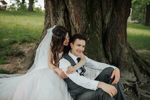 ein schön Braut mit lange lockig Haar im ein schick Kleid Umarmungen das Bräutigam, lächelt, suchen in das Linse unter ein groß Baum. Porträt von das Braut und Bräutigam. Frühling Hochzeit. natürlich bilden foto