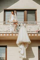 Weiß Hochzeit Kleid hängend auf Balkon Geländer, breit Schuss von Haus im Vorderseite und Mädchen auf Balkon. Hochzeit Einzelheiten, modern Hochzeit Kleid mit lange Zug, lange Ärmel und öffnen zurück foto