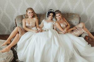 ein Brünette Braut im ein Weiß elegant Kleid und Krone und ihr blond freunde im golden Kleider sind Sitzung auf das Sofa im das Braut Zimmer, lächelnd und posieren. Hochzeit Porträt, Hochzeit Foto