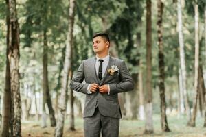 ein stilvoll Bräutigam Spaziergänge nach vorne im das Wald Knöpfen seine Jacke zwischen hoch Bäume. ein modisch Bräutigam, ein Erwachsene Mann foto