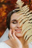 ein Mädchen mit ein schön Frisur und bilden ist Stehen im ein Weiß Hochzeit Kleid im ein Herbst Wald auf ein Hintergrund von Orange Blätter, halten ein Farn Ast im ihr Hände foto