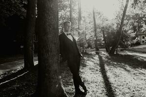 Porträt von das Bräutigam im das Wald im ein breit Schuss, das Bräutigam lehnt sich seine Schulter gegen ein Baum, das Foto ist hinterleuchtet. schwarz und Weiß Foto